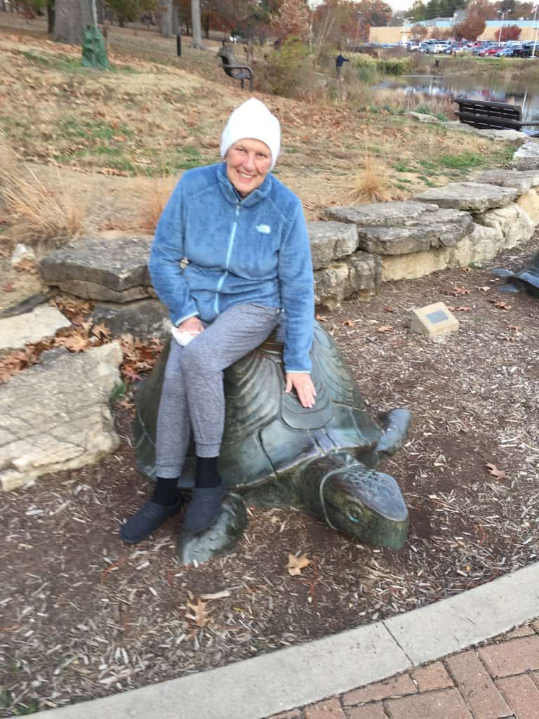 Beth Purcell, smiling and sitting on a turtle at Walker Lake in Kirkwood, MO, after losing her hair to chemo.  